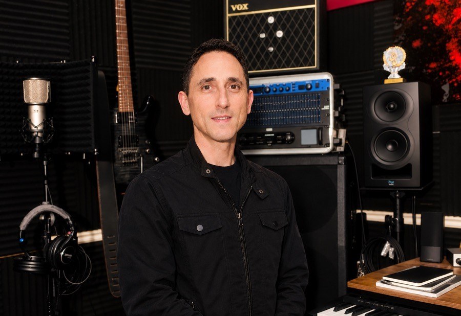 Theron Rodriguez, founder of The Concert Consulting Group, standing in his home studio surrounded by a guitar, microphones, studio monitors, and rack gear